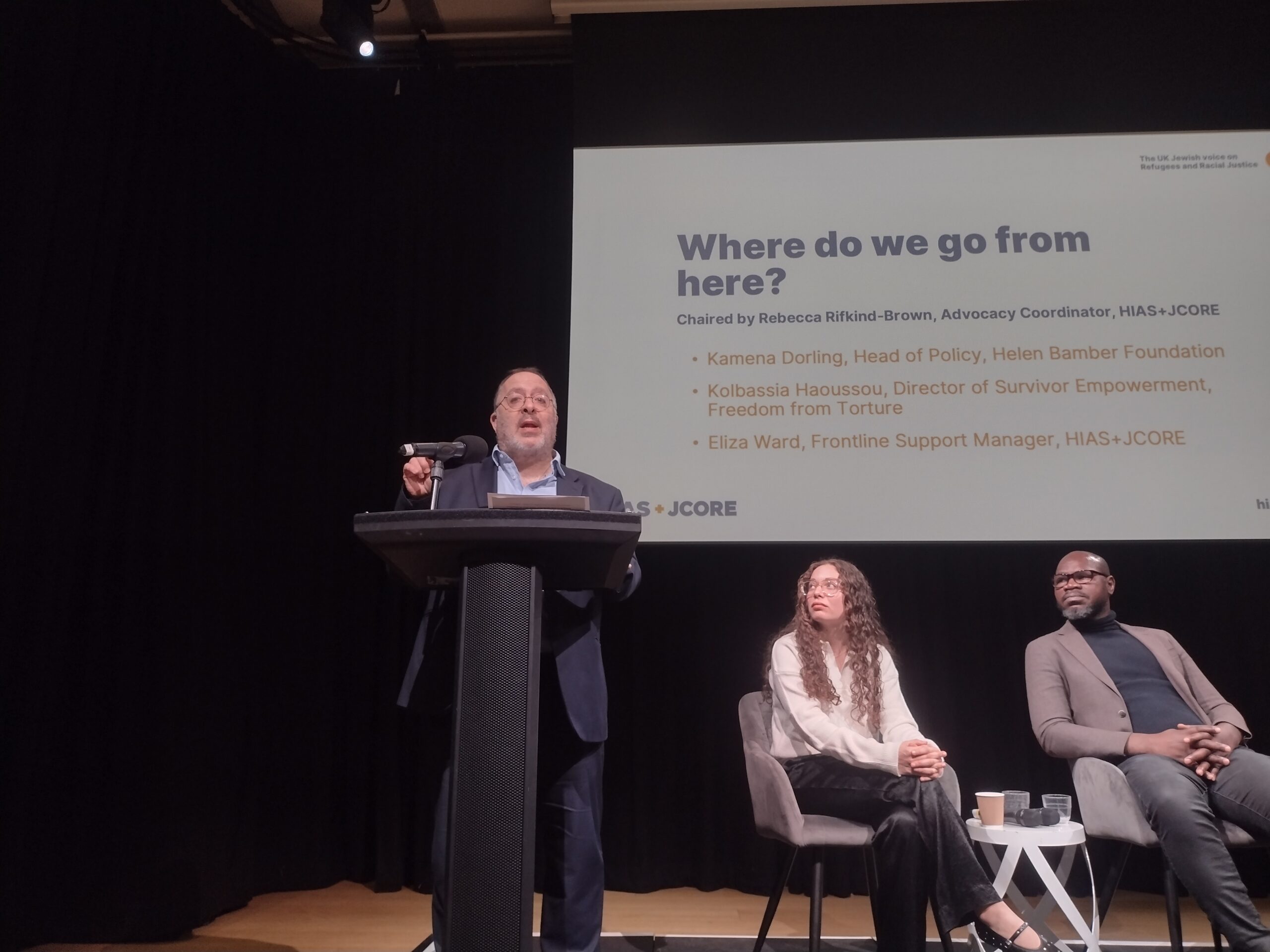 Rabbi David Mason addresses the audience, stood at a lectern. To his right are Rebecca Rifkind-Brown and Kolbassia Haoussou MBE.
