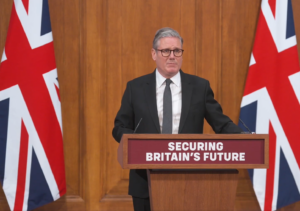 Sir Keir Starmer stands at a lectern. To his left and right are large Union Jack flags.